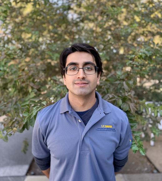 Young man with glasses wearing gray quarter-zip with chest logo, standing outdoors by leafy tree