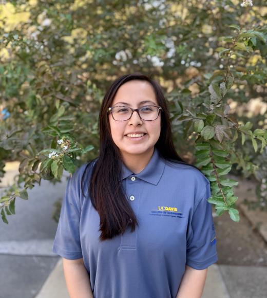 Smiling young woman with glasses wearing blue polo, standing before leafy green shrub