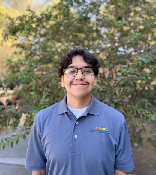 Smiling young person with curly dark hair and glasses wearing blue polo, green outdoor background