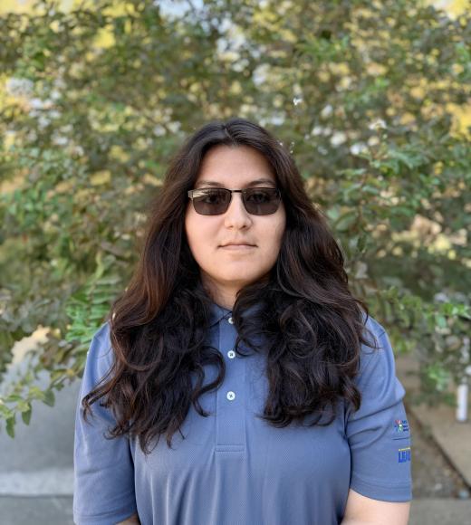 Young woman with long dark hair, sunglasses, blue polo, leafy outdoor background (photograph)