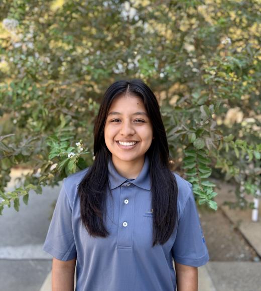Smiling young woman with long dark hair in blue polo, leafy background, photograph