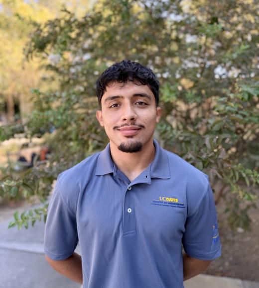 Young man with slight smile in blue polo, standing outdoors with blurred leafy background