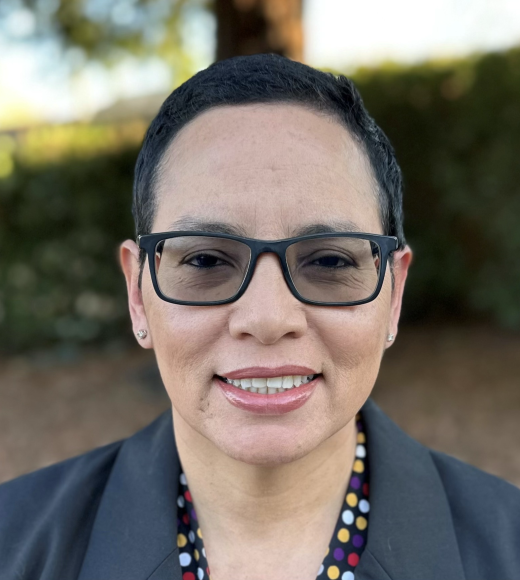 Close-up portrait of a smiling person with short dark hair, glasses, blazer outdoors