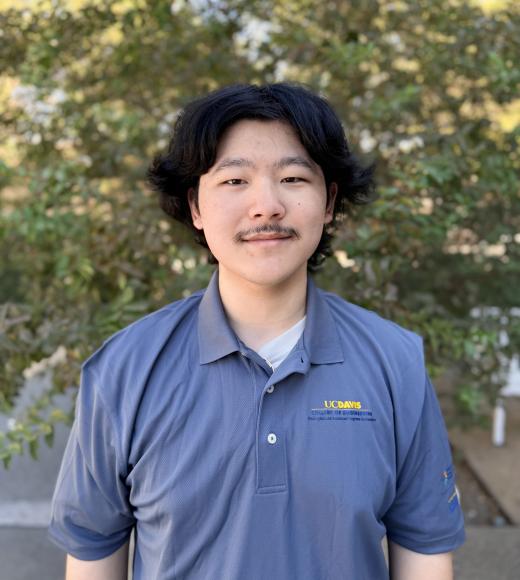 Headshot photo of young man with mustache in blue polo, leafy background