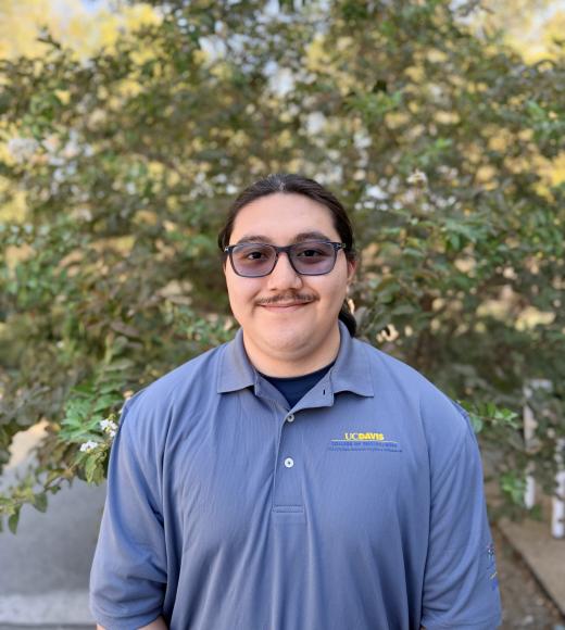 Young person with glasses and mustache, blue polo, smiling in front of leafy tree