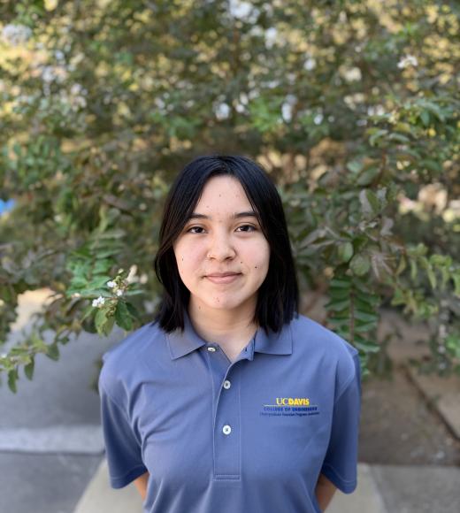 Young woman in blue polo with embroidered logo, smiling, green foliage background