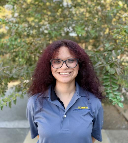 Smiling young woman with curly dark red hair and glasses in gray polo, leafy background