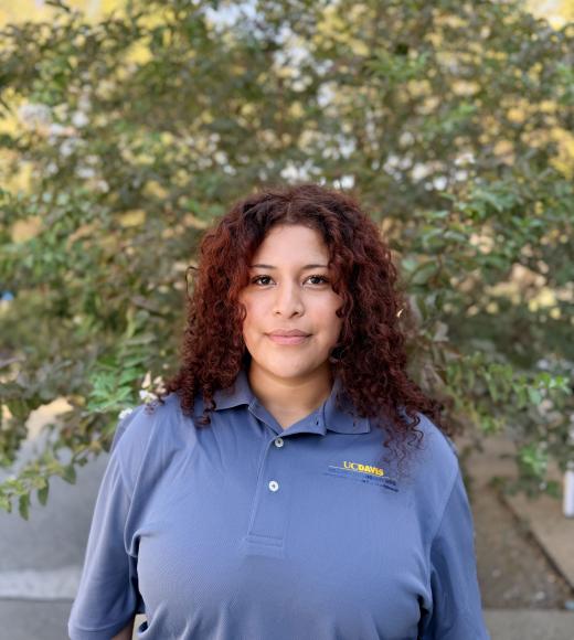 Person with shoulder-length curly auburn hair in blue polo, trees behind