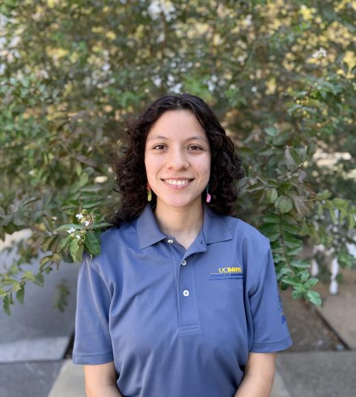Smiling young woman with dark curly hair in blue polo, standing before a leafy tree