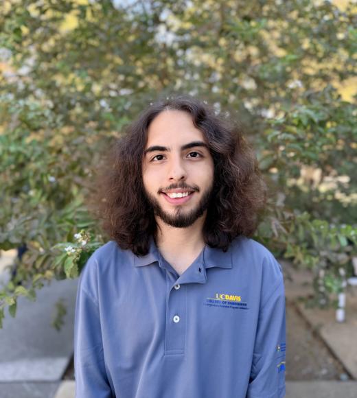 Casual headshot photo of smiling young man with curly hair, beard, and blue polo, trees behind