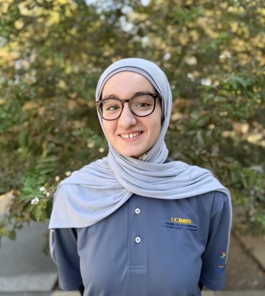 Smiling young woman in light blue headscarf and glasses, navy polo, trees blurred behind