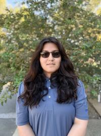 Young woman with long dark hair, sunglasses, blue polo, leafy outdoor background (photograph)