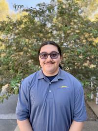 Young person with glasses and mustache, blue polo, smiling in front of leafy tree